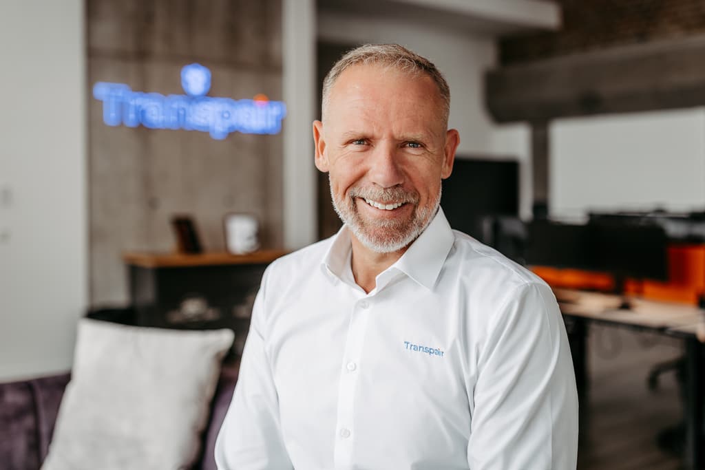 Reinhard Heesen, Geschäftsführer von Transpair, lächelnd im Büro vor dem Transpair-Logo